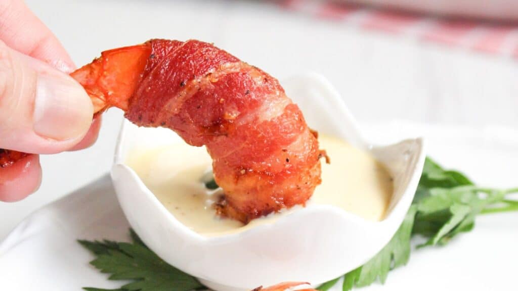 A hand holding a bacon-wrapped shrimp dipped in a small bowl of sauce, with a green leaf garnish on the side.