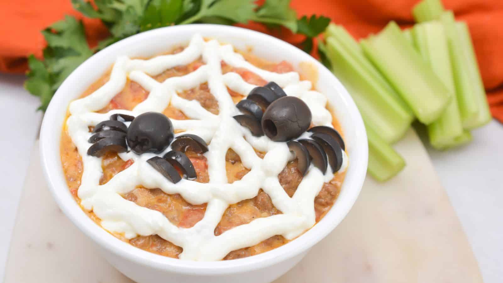 A bowl of beef queso dip with a spider web on top served with celery sticks.