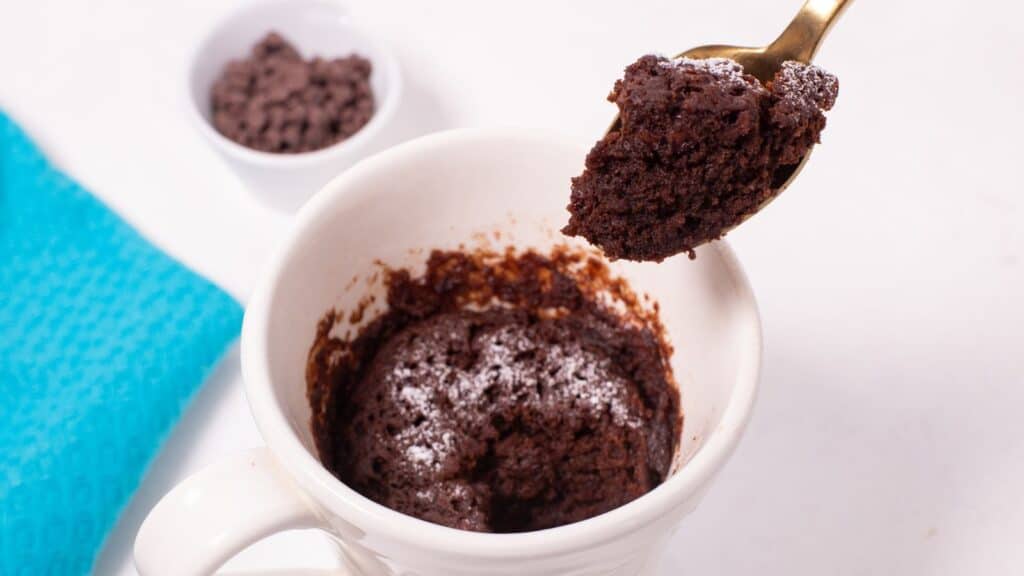 A spoon lifting a bite of freshly made chocolate mug cake from a white mug, with chocolate chips in a small bowl in the background.