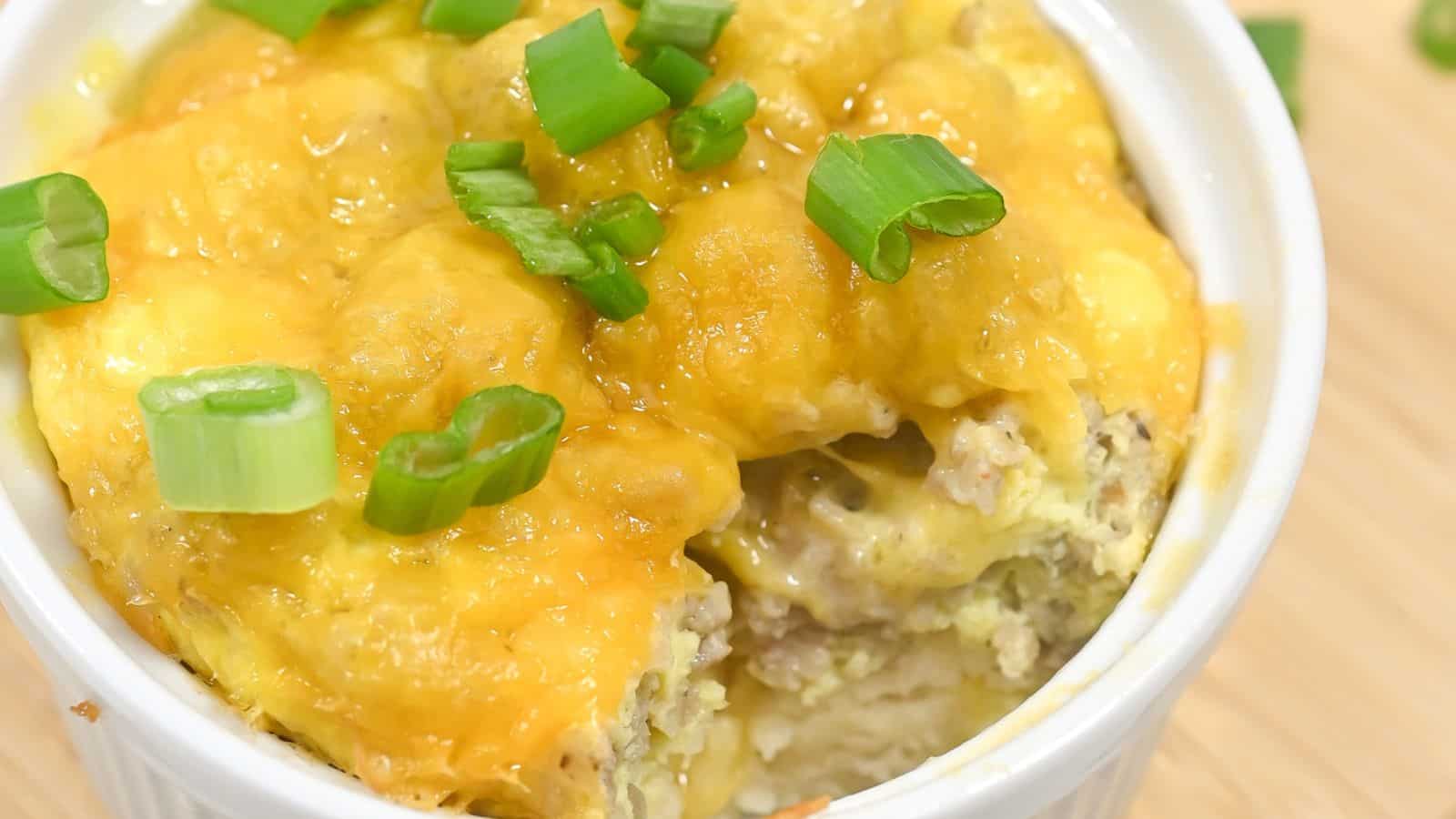 A white bowl filled with a cheesy, layered casserole topped with melted cheddar cheese and chopped green onions.