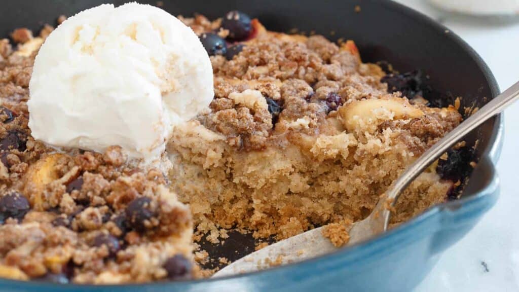 A close-up image of a scoop of vanilla ice cream on top of a freshly baked fruit cobbler in a blue pan, with a spoon on the side.