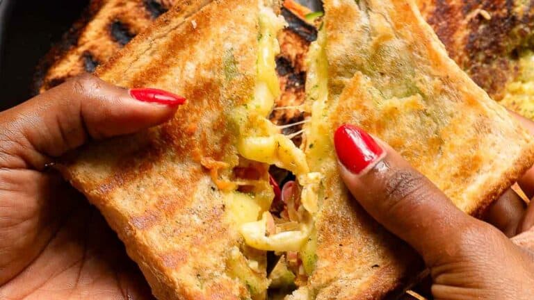 Hands with red nail polish holding a grilled cheese sandwich, split open to show melted cheese and fillings.