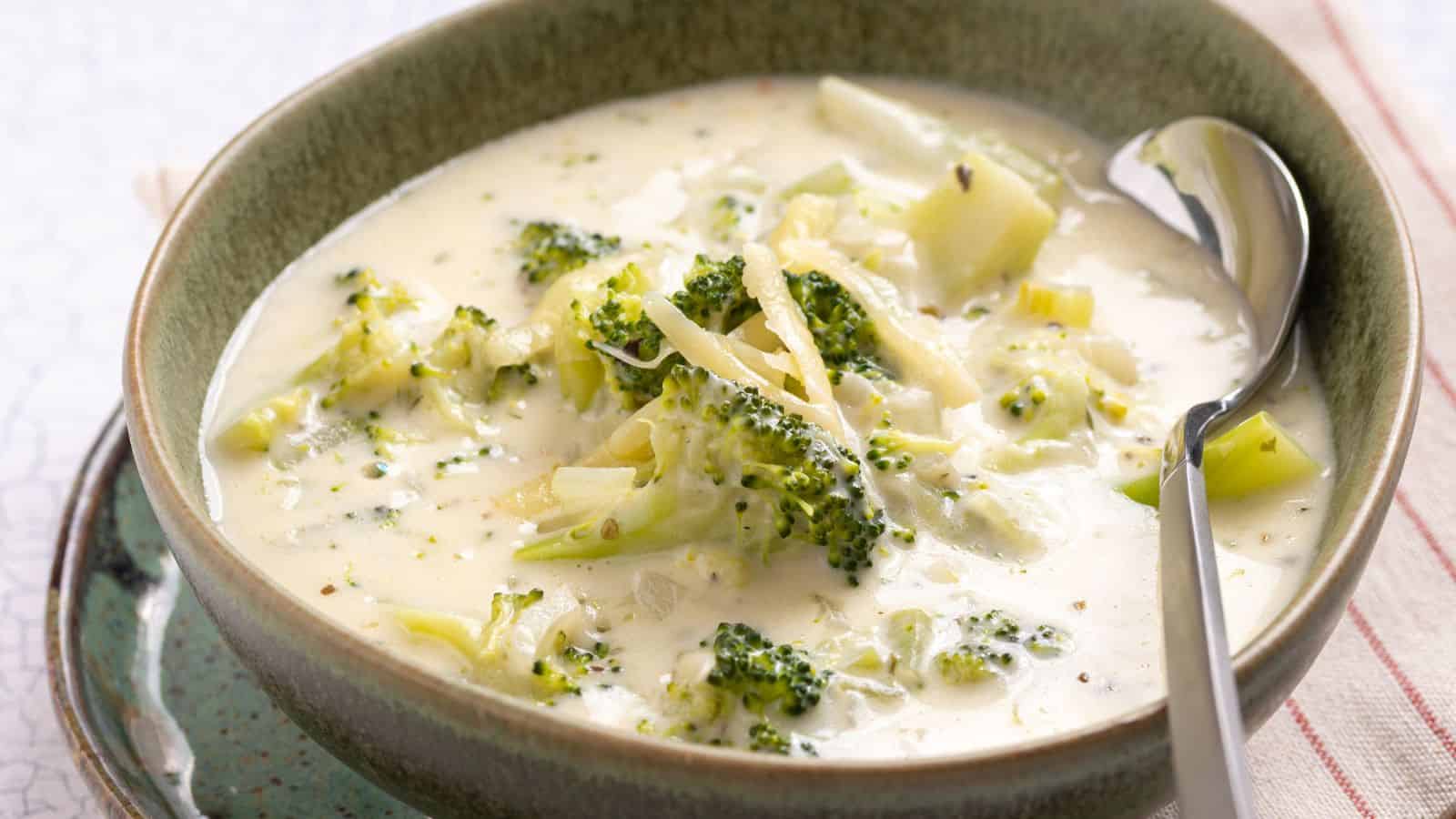 A bowl of creamy broccoli cheese soup beside fresh broccoli and shredded cheese, with a slow cooker in the background.