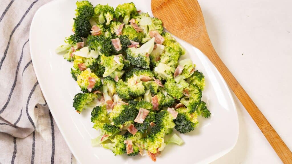 A plate of broccoli salad with bacon bits, served on a white oval dish with a wooden spoon, on a kitchen towel.