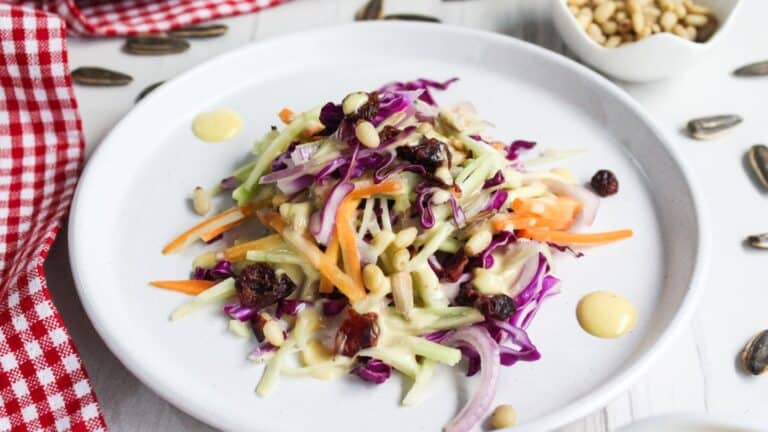 A plate of colorful broccoli slaw with shredded purple cabbage and carrots topped with dried cranberries and pine nuts, next to a red-checked napkin.