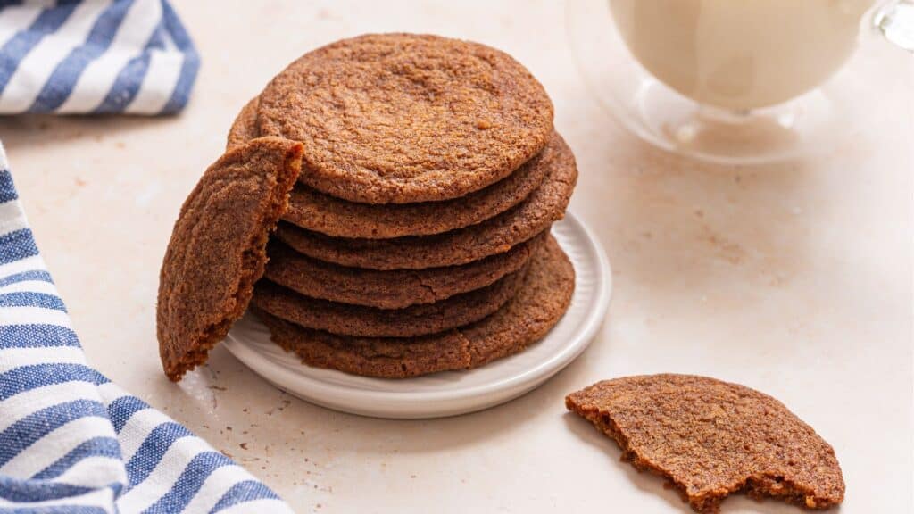 A stack of small-batch crispy brown cookies on a white plate with a glass of milk in the background and a striped napkin to the side.