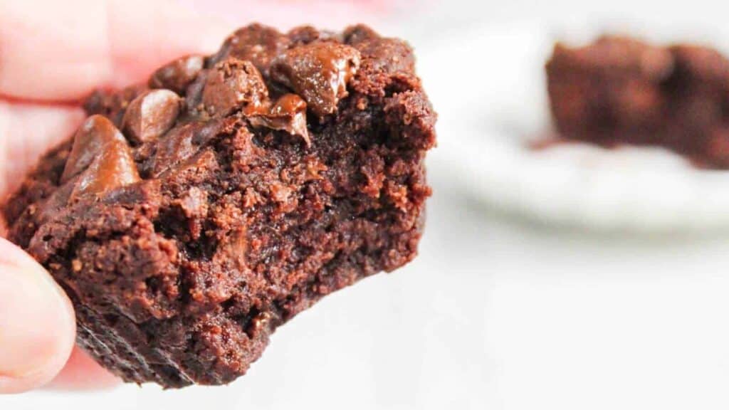 Close-up of a hand holding a half-eaten chocolate brownie with gooey chocolate chunks, with another partial brownie blurred in the background.