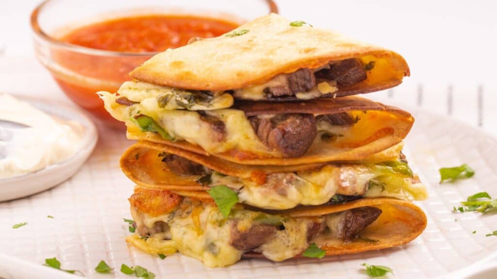A stack of quesadillas filled with cheese and meat, served with a side of red salsa on a white plate.