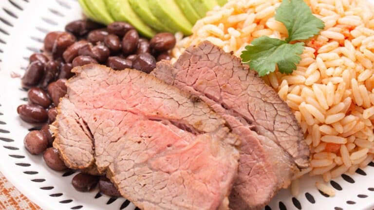 A plate of sliced carne asada with sides of white rice, black beans, and sliced avocado, garnished with a sprig of parsley.