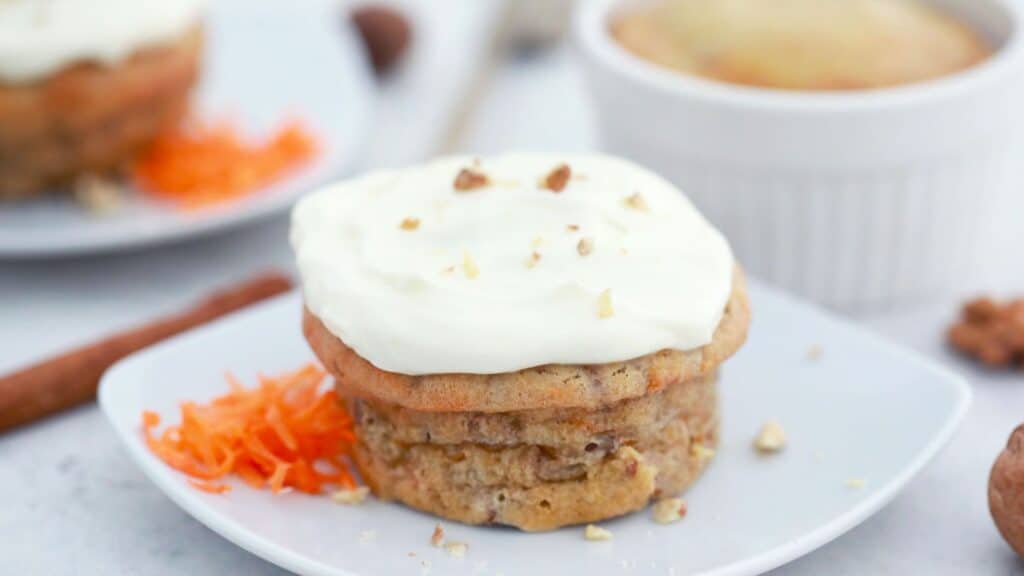 Carrot cake muffin topped with cream cheese frosting and chopped nuts, served on a white plate with grated carrot decoration.