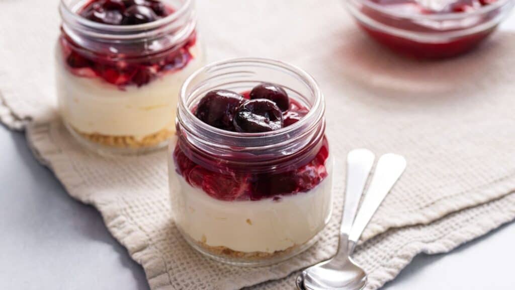 Two jars of layered cheesecake with a graham cracker base, creamy filling, and topped with cherry compote, accompanied by spoons on a cloth.