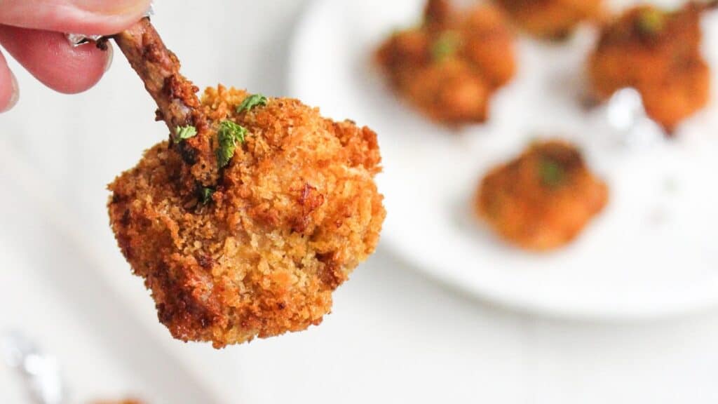 A close-up of a hand holding a crispy breaded chicken drumstick over a plate of similar drumsticks garnished with parsley.