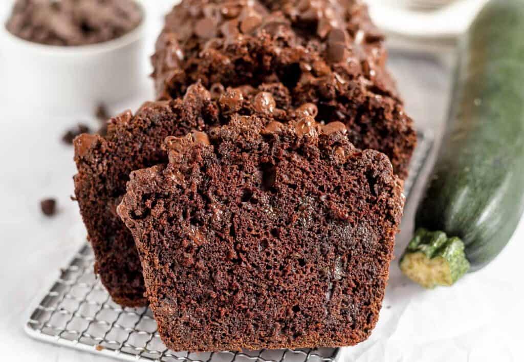 Slices of rich chocolate zucchini bread with chocolate chips on a wire cooling rack, with a whole zucchini and extra chocolate in the background.