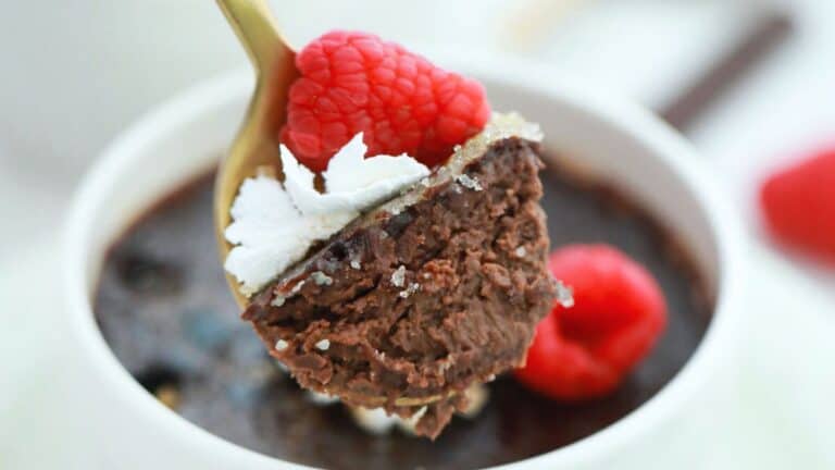 A spoonful of chocolate creme brulee garnished with a raspberry and whipped cream, above a white bowl containing more dessert.