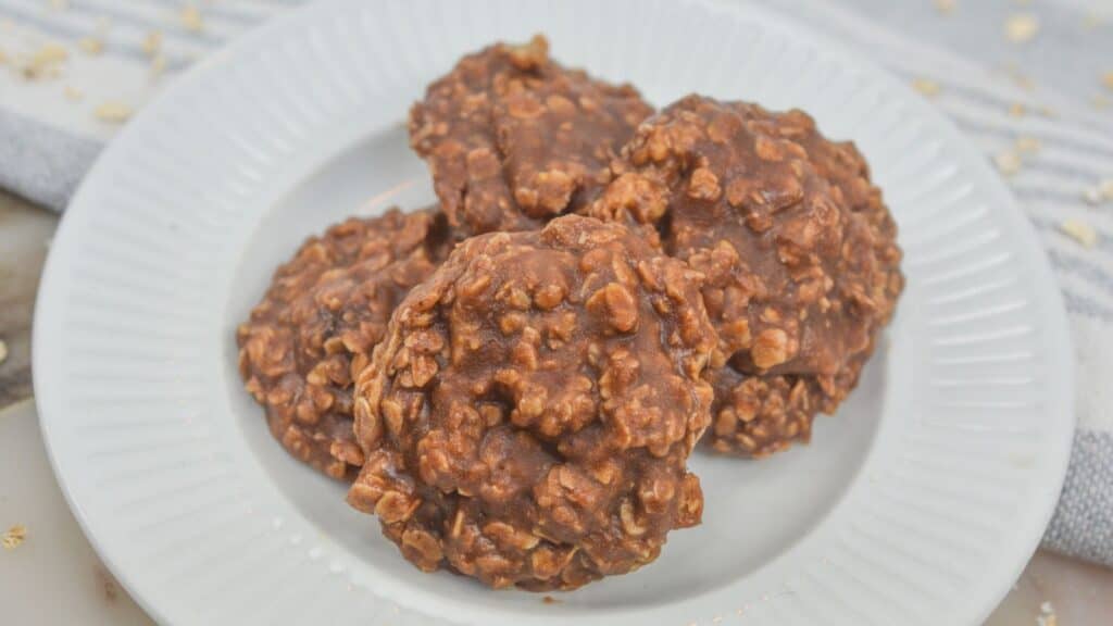 Three no-bake chocolate oatmeal cookies on a white plate, topped with visible oats and crumbs on a light-colored tabletop.