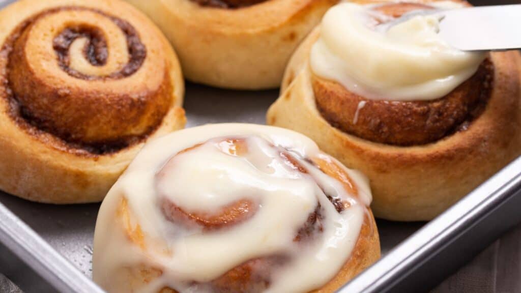 A knife spreading cream cheese frosting on freshly baked cinnamon rolls in a metal tray.