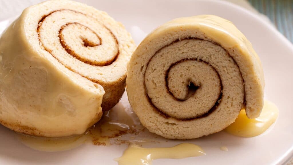 Two slices of cinnamon roll cake covered in creamy frosting, placed on a white plate with extra drizzle of sauce.