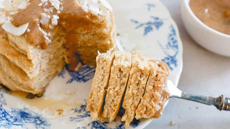 A slice of layered peanut butter cake on a decorative plate with a fork, topped with drizzle and coconut flakes.