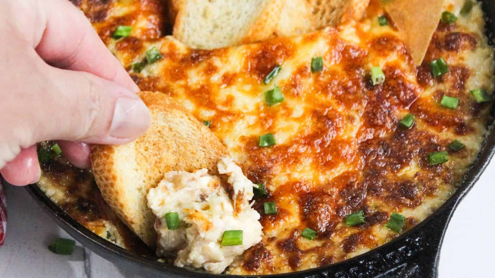 A person dipping a piece of bread into a skillet of cheesy dip.