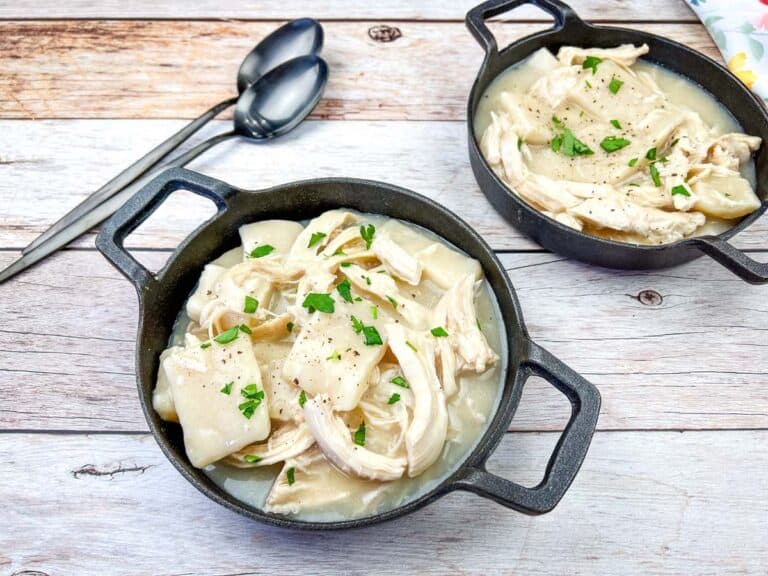 Two skillets of chicken and dumplings garnished with parsley, with spoons on a wooden table.