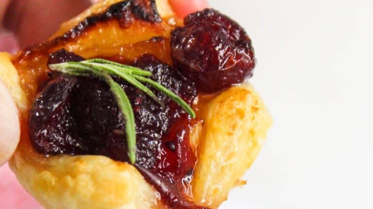 Close-up of a puff pastry topped with cranberries and a sprig of rosemary, held against a white background.