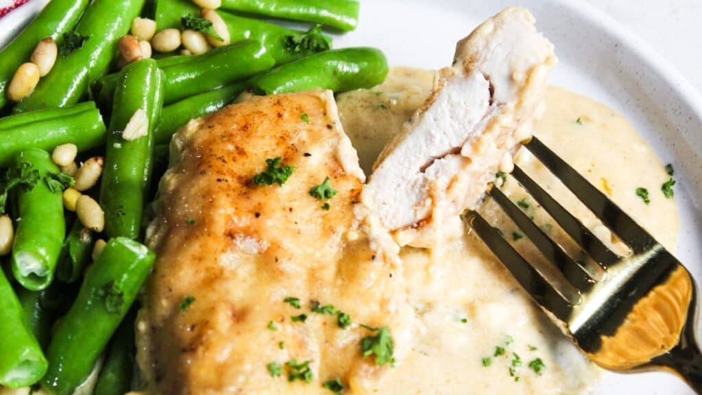 A plate with a slice of chicken breast, green beans, and pine nuts with a fork resting on the chicken.