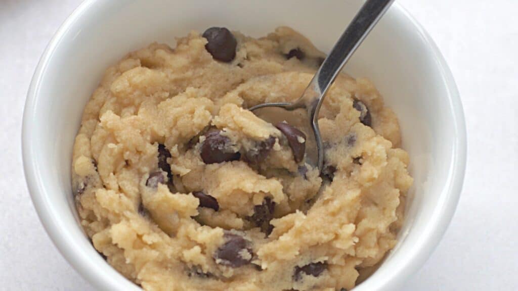 Bowl of raw cookie dough with chocolate chips and a spoon, on a white background.
