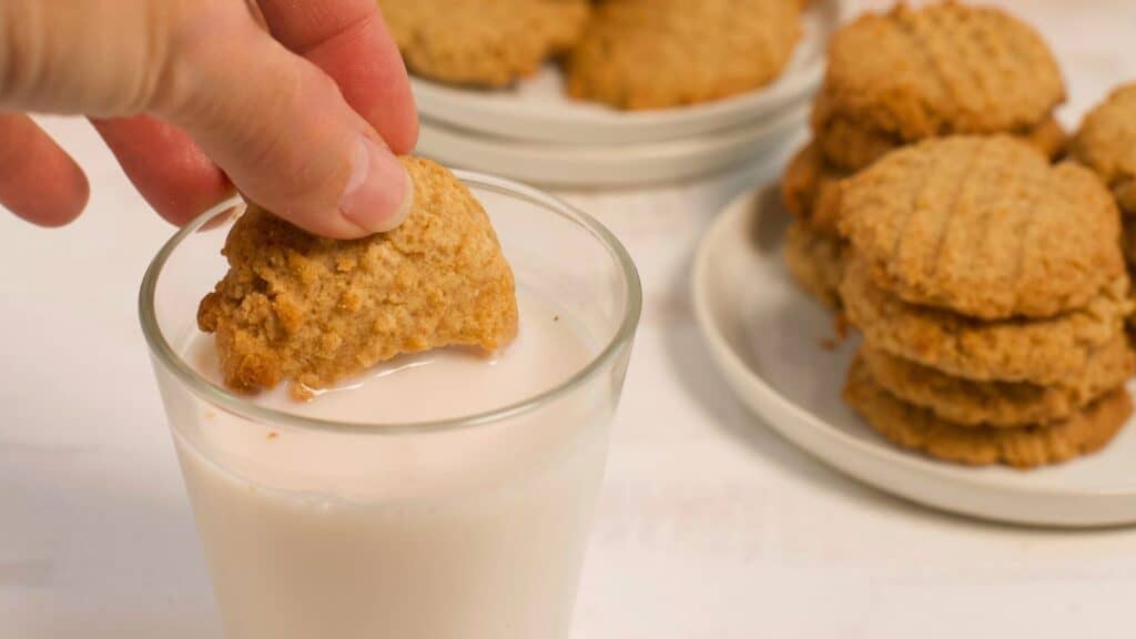 A person dipping a peanut butter cookie into a glass of milk, with more cookies stacked on a plate in the background.