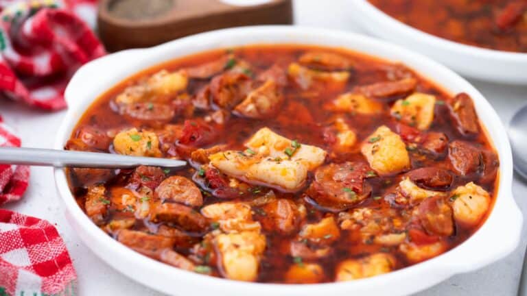 A bowl of spicy seafood stew with shrimp, sausage, and chunks of fish, garnished with herbs, served with a spoon on a table.