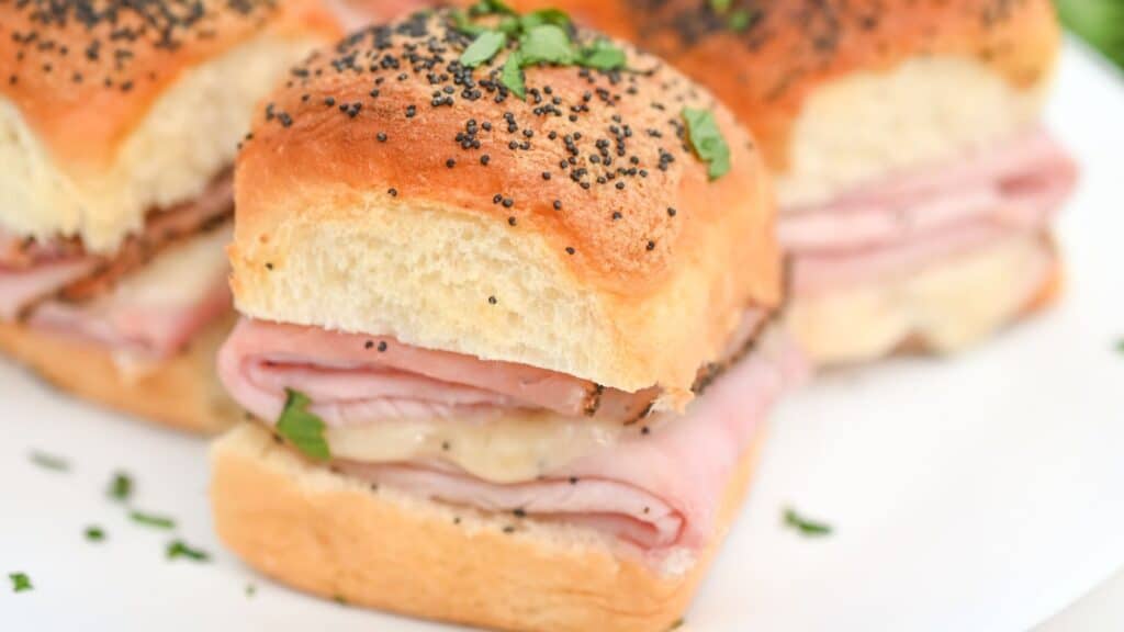 Slide of ham and cheese sandwiches on poppy seed rolls garnished with chopped herbs, displayed on a white plate.