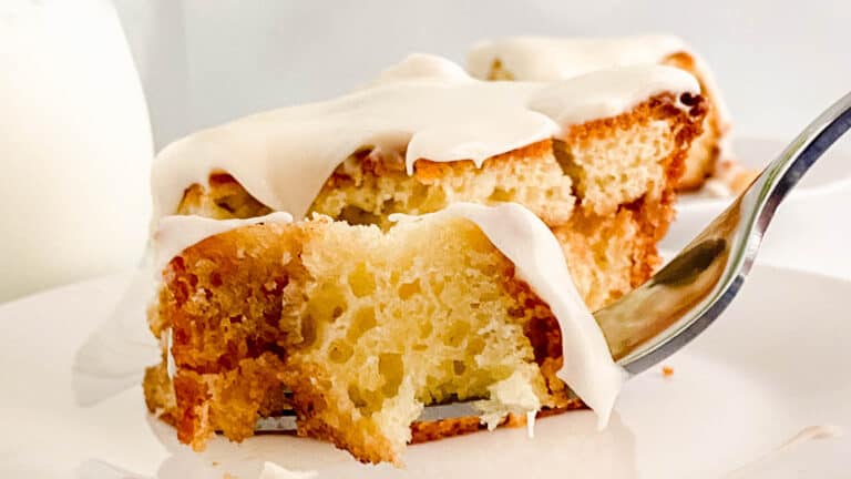 A close-up image of a slice of cinnamon cake with creamy white frosting, a fork is lifting a bite from the plate.