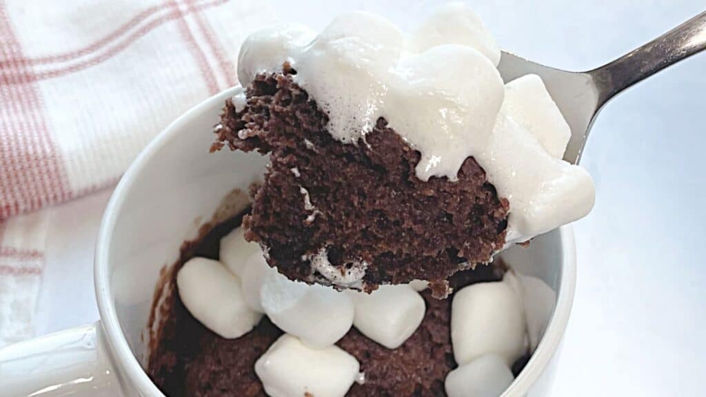 A spoon lifting a bite of chocolate mug cake topped with melted marshmallows from a white mug.