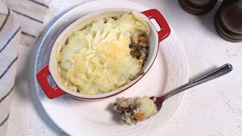 Shepherd's pie in a red ceramic dish with a spoon, showcasing layers of mashed potatoes and minced meat with vegetables on a white tablecloth.