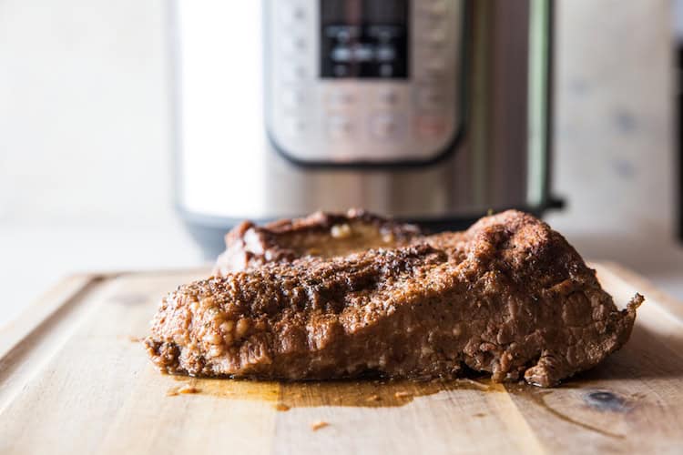 A cooked steak rests on a wooden cutting board in front of a modern electric pressure cooker on a kitchen counter.