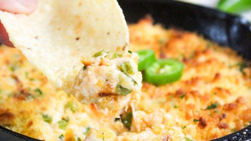 A close-up of a hand dipping a tortilla chip into a skillet of bubbling, cheesy jalapeño popper dip.