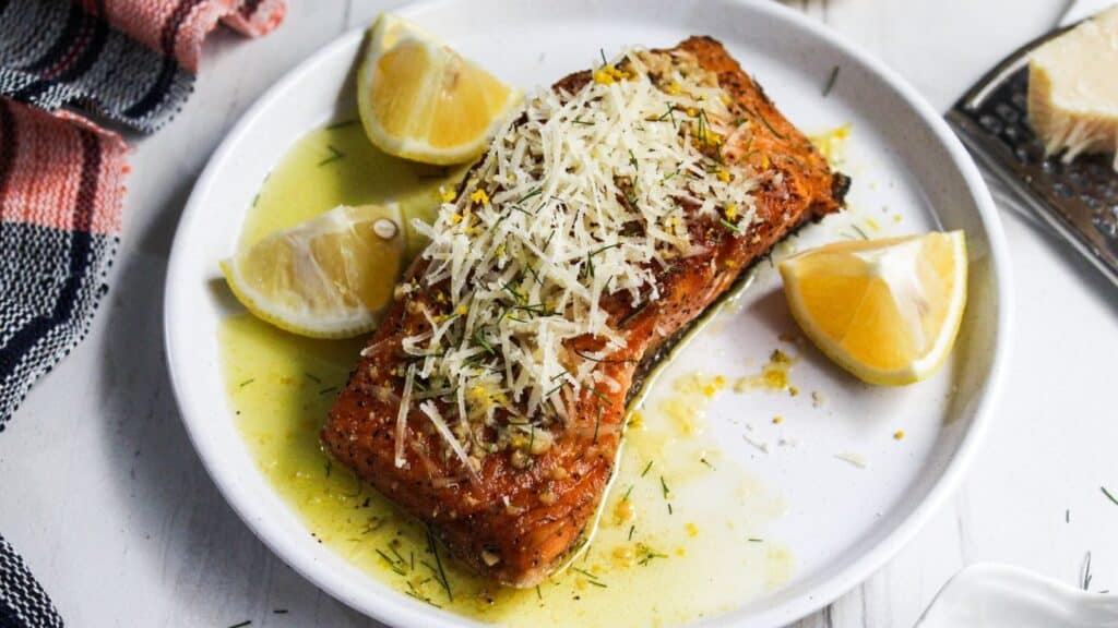 A grilled salmon fillet garnished with grated cheese and herbs, served with lemon wedges on a white plate.
