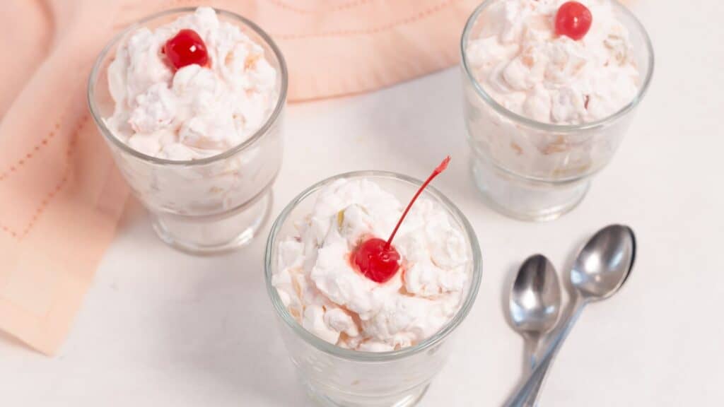 Three glasses filled with marshmallow fruit salad and topped with red cherries on a light pink cloth, accompanied by two spoons.