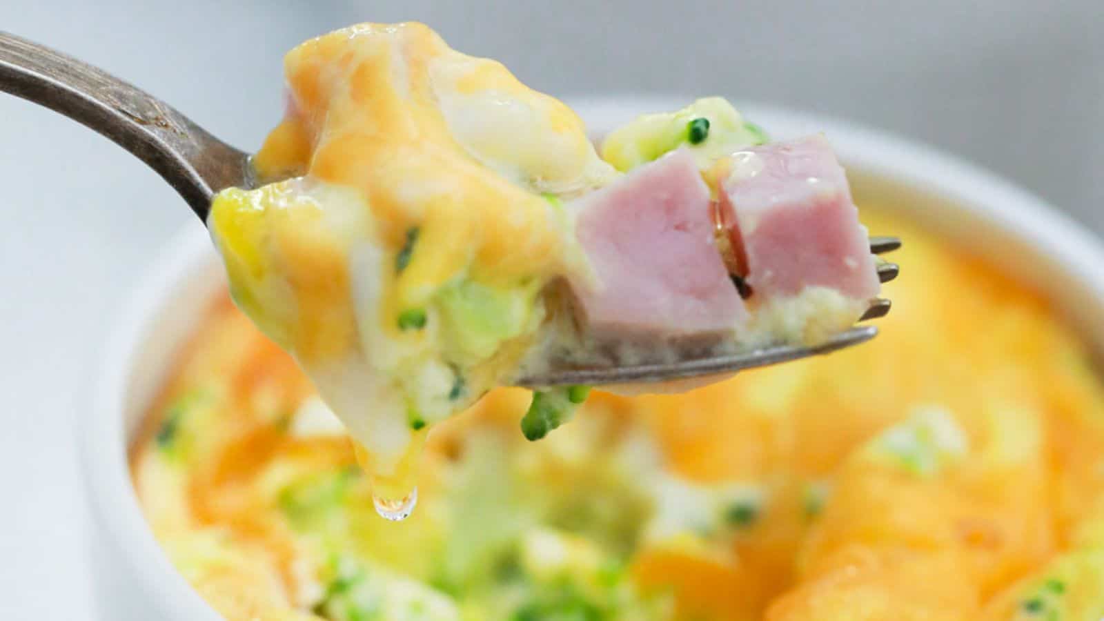 A fork holding a bite of mini frittata with ham and broccoli, with the dish in the background.