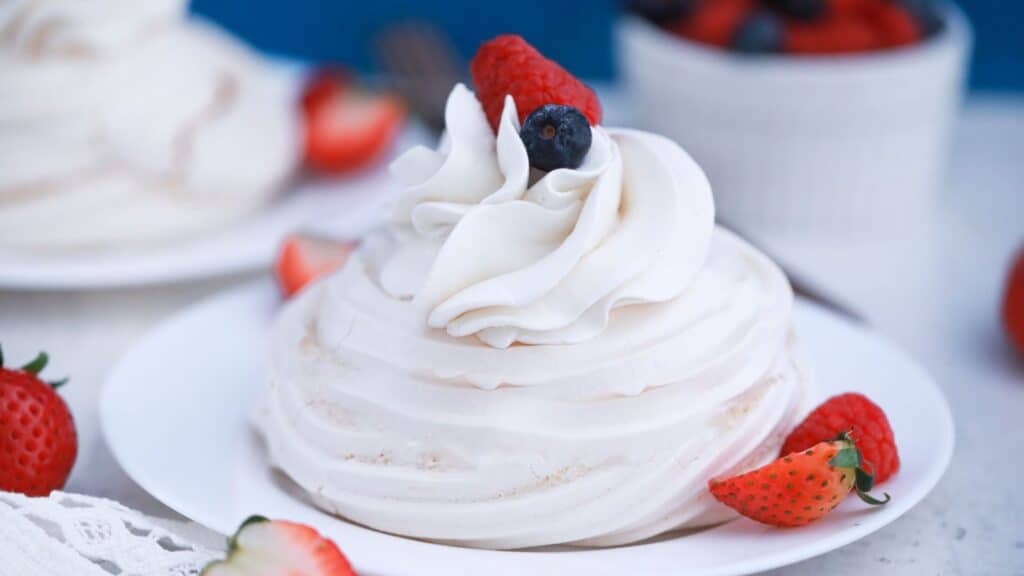 A pavlova dessert with whipped cream and berries on a white plate, surrounded by fresh strawberries.
