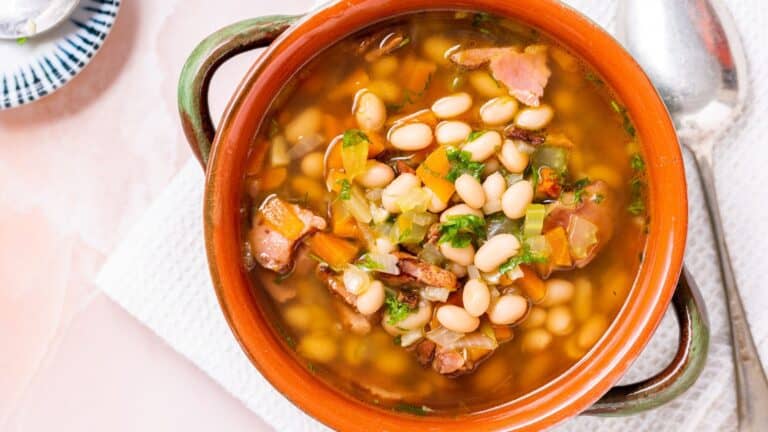 A bowl of bean soup with chunks of ham and diced vegetables, served in a rustic terracotta bowl with a spoon on the side.