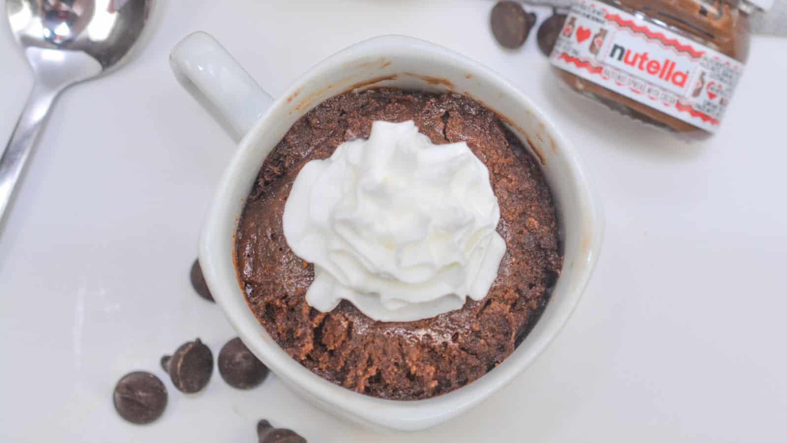 Homemade chocolate mug cake topped with whipped cream, served with a spoon and a small nutella jar on the side.