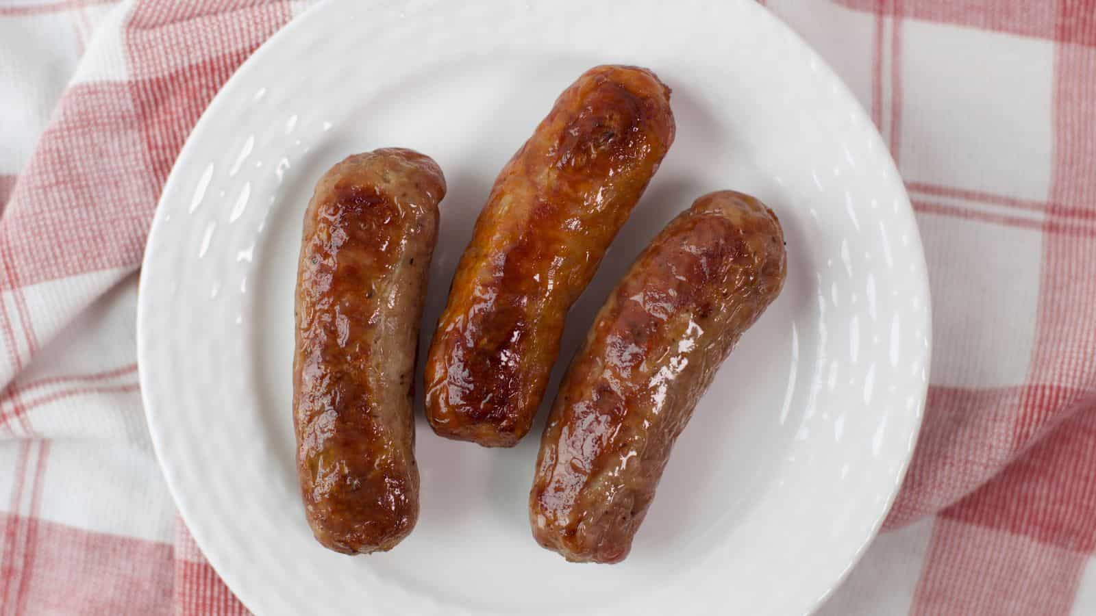 Three oven-baked sausages on a white plate with a red and white checkered napkin in the background.