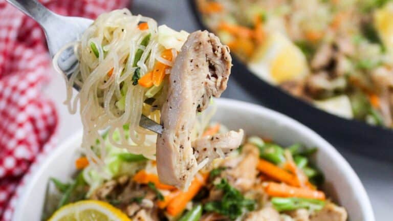 A fork lifting a bite of chicken and noodles with vegetable stir-fry from a bowl.