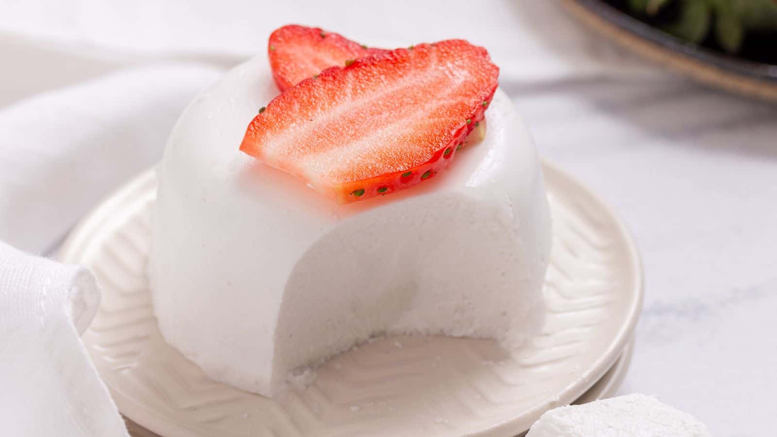 A slice of white, creamy panna cotta topped with a fresh strawberry slice on a white plate, with a textured napkin beside it.