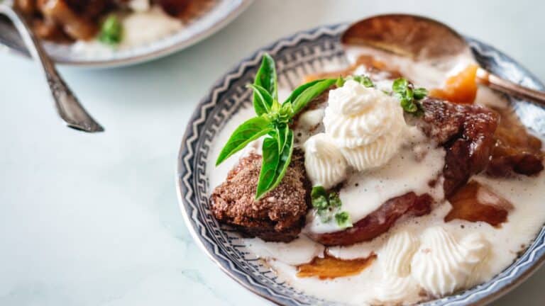 A plated dessert featuring a slice of peach pie topped with whipped cream and fresh basil leaves, served with a spoon.