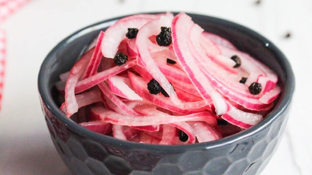 A bowl of pickled red onions garnished with black peppercorns, placed on a white surface.