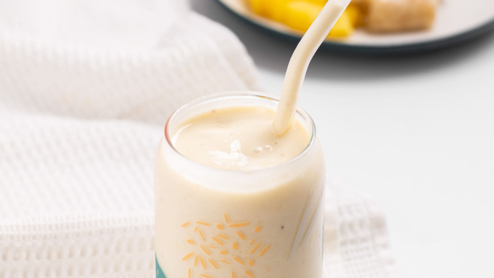 A close-up of a pineapple banana smoothie in a glass with a straw, surrounded by a white napkin with pineapple and banana in the background.