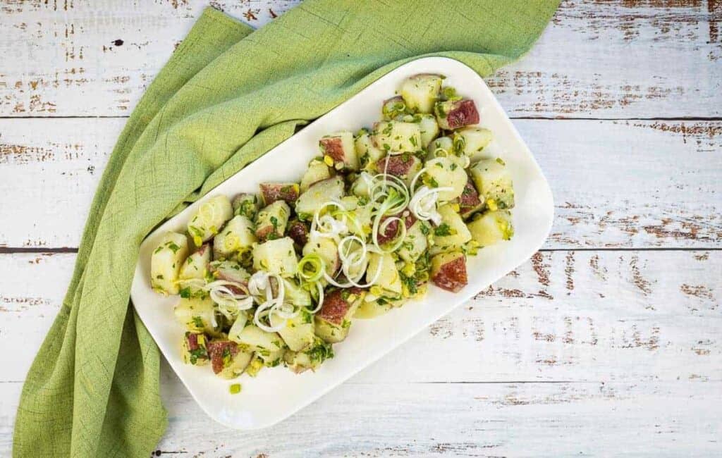 A plate of cucumber salad with diced red potatoes and sliced onions, garnished with herbs on a white wooden table with a green napkin.