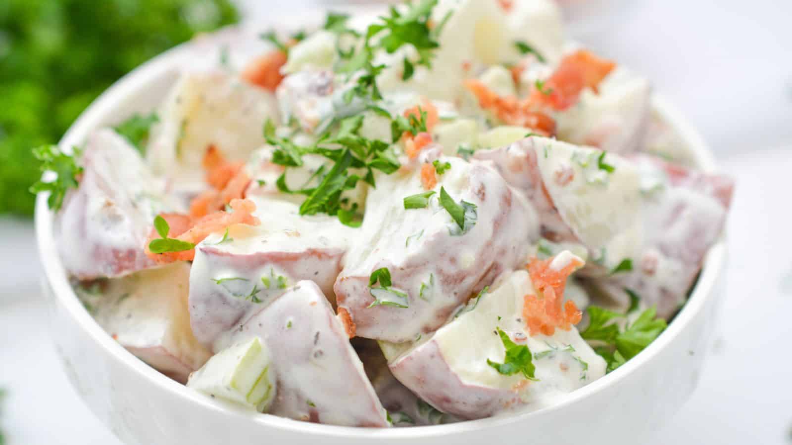 Bowl of potato salad garnished with fresh herbs and bacon on a white background.