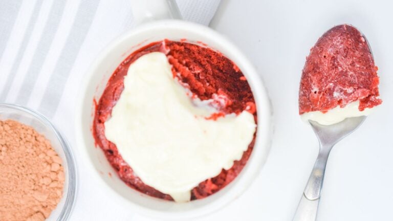 Top view of a red velvet cake with a creamy frosting in a white bowl, next to a spoon and a small container of cocoa powder.